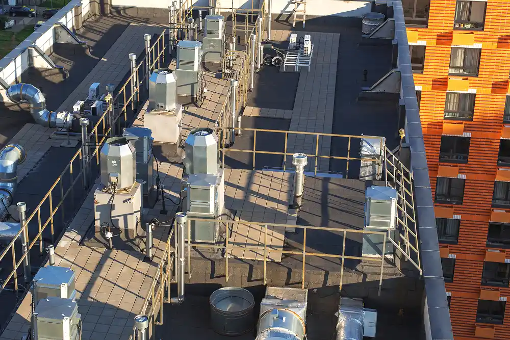 Rooftop view with numerous metal HVAC units and ventilation pipes, surrounded by safety railings. Adjacent building features a bright orange facade.