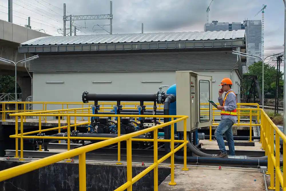A worker in a hard hat and safety vest inspects an industrial control panel near large pipes, set against a backdrop of urban construction and cloudy skies.