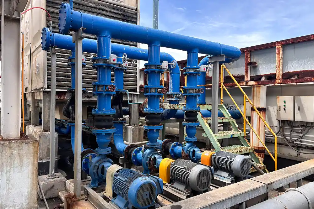 A series of blue industrial pumps and pipes are connected in a mechanical setup. The setting is outdoors, with visible stairs and a control panel, under a clear sky.