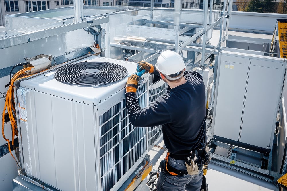 Worker in protective gear fixes a rooftop air conditioning unit using a drill. The scene is industrial, conveying focus and technical expertise.